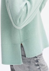 Pull bleu clair en tricot avec une fente latérale à l'ourlet, porté sur un jean denim gris, montrant la texture du tissu et les détails des coutures.