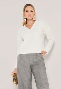 Vrouw in een wit gebreid sweater, grijze geblokte broek, grote zilveren en gouden driehoekige oorbellen, met een gouden clutch handtas.