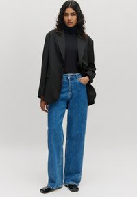 Zwarte oversized blazer over een zwarte coltrui, gecombineerd met hoge taille wijde blauwe jeans en zwarte schoenen, op een neutrale grijze achtergrond.
