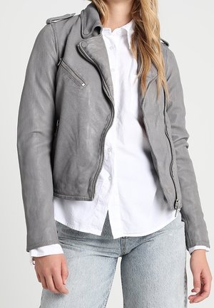 Vrouw draagt een grijze leren motorjack over een wit blousehemd en lichtblauwe spijkerbroek, staand tegen een effen achtergrond.