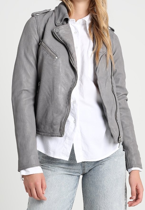Femme portant une veste de motard en cuir gris par-dessus une chemise blanche à boutons et un jean bleu clair, debout devant un fond uni.