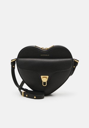 Sac besace en cuir noir en forme de cœur avec fermoir doré, bandoulière ajustable, et nom de la marque "Coccinelle" embossé à l'avant.