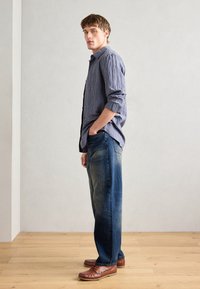 Junger Mann, der seitlich steht, mit den Händen in den Taschen, trägt ein blau kariertes Hemd, faded blaue Jeans und braune Lederloafer auf einem Holzboden.