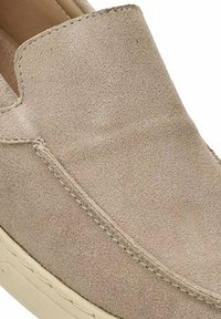 Zapato de ante beige de tipo slip-on con puntera redondeada, detalle de costura mínima y suela de goma de color claro. Textura suave y diseño de perfil bajo.