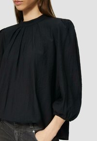 Schwarze, leichte Bluse mit hohem Kragen und langen, gepufften Ärmeln mit gerafften Details, die ein strukturiertes Erscheinungsbild schaffen.
