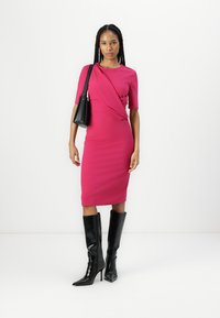 Robe midi fuchsia à manches courtes avec un détail de design tordu, associée à des bottes à talons aiguilles noires jusqu'aux genoux et un petit sac à main noir.