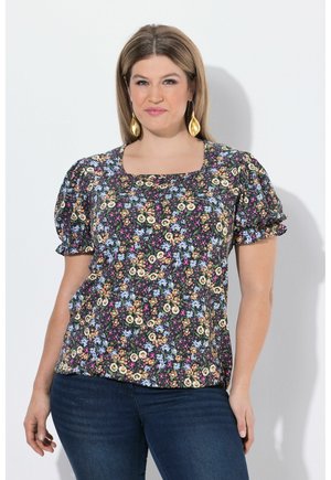 Frau in einer schwarzen Bluse mit Puffärmeln und floralem Muster sowie dunkelblauer Jeans, die vor einem weißen Hintergrund steht.