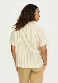 Femeie cu păr creț, purtând o cămașă bej cu mânecă scurtă, cu dungi verticale, și pantaloni culoarea bronzului, văzută din spate pe un fundal simplu.