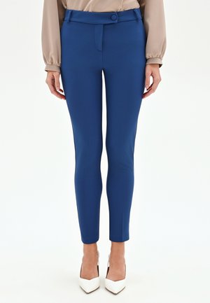 Donna che indossa pantaloni aderenti blu alla caviglia con vita alta, chiusura con bottoni, abbinati a una camicia beige e décolleté bianche con punta affilata.