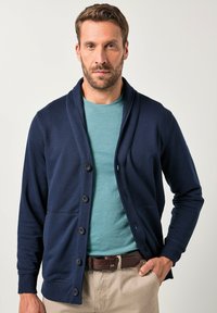 Námořnický pletený kardigan s šálovým límcem, šesti knoflíky a dvěma kapsami, nahoře na světle modrém tričku, nošený s khaki kalhotami.