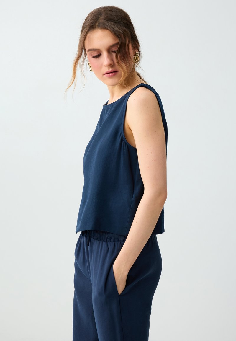 Femme aux cheveux ondulés portant des boucles d'oreilles en or, un haut sans manches bleu marine et un pantalon bleu marine, les mains dans les poches, regardant pensivement vers le bas.