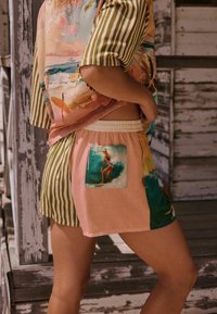 Personne portant un short et une chemise à manches courtes avec des motifs mélangés, comprenant des rayures, de l'art abstrait et une scène de surf sur la poche.