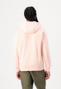 Sweatshirt com capuz de cor rosa claro, feito de tecido suave, com um bolso canguru e punhos canelados, combinado com calças verdes-oliva.