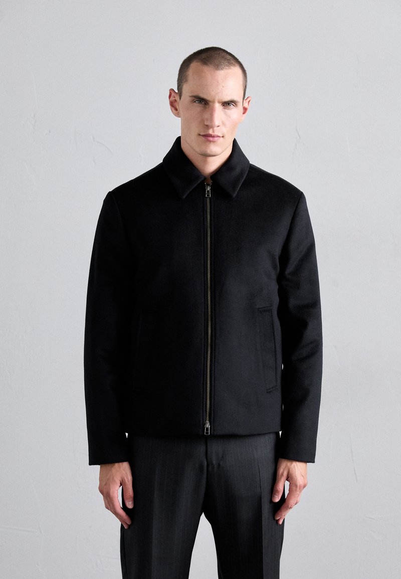 Black zippered jacket with a collar, made of smooth fabric. Features side pockets and a tailored fit. Worn over gray pinstriped pants.