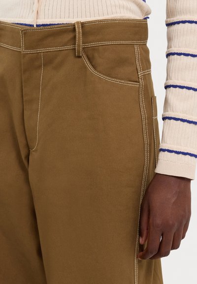 Pantalones de algodón marrón con detalles de costura blanca, que presentan un bolsillo frontal y un diseño de pierna recta, exhibidos junto a un suéter a rayas.