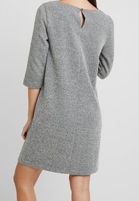 Robe en maille grise avec manches trois-quarts, une ouverture en forme de goutte d'eau au dos, et un motif texturé. Coupe simple et droite.