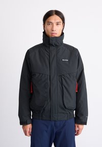 Sort Columbia-jakke med høj krave, frontlynlåse, to store lommer, røde detaljer og elastiske manchetter. Letvægts, isoleret design.