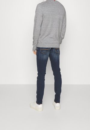 Homme portant un pull gris, un jean bleu foncé et des baskets blanches, debout face à un mur blanc, les mains détendues.