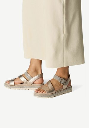 Metallic bronze sandals feature wide straps and a textured ankle strap with buckle, set on a cushioned sole with a rugged tread.