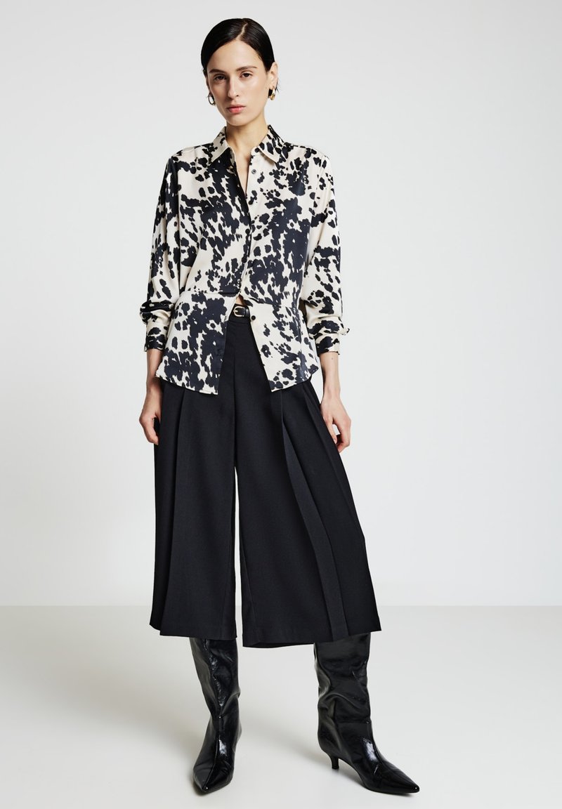 Donna in piedi che indossa una camicia a bottoni bianca con macchie nere, culotte nere a gamba larga e stivali neri alti fino al ginocchio con punta affilata.