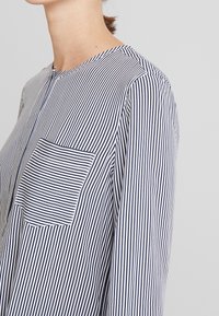 Blusa a righe blu navy e bianca con scollo tondo, maniche lunghe e taschino sul petto con righe verticali, realizzata in tessuto leggero.
