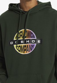 Zelená mikina s kapucí vyrobená z bavlněné směsi, s kulatým, barevným logem, které obsahuje text "WORLDWIDE COMPANY" a "DO SHOE" v blokových písmenech.