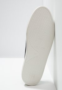 Semelle d'une basket noire avec une semelle extérieure en caoutchouc blanche à motif, taille marquée 37, positionnée verticalement contre un mur blanc.