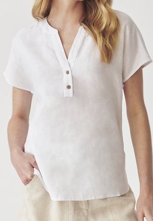 Femme portant une blouse en lin blanche à manches courtes avec deux boutons marron et un pantalon beige, main posée négligemment dans la poche.