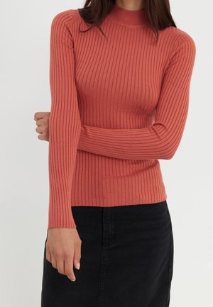 Kvinde iført en tætsiddende, langærmet, ribbet rust-orange sweater og en sort nederdel, stående med den ene arm krydset over kroppen.