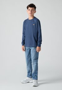 Ναυτικό μπλουζάκι με μακρύ μανίκι και μικρό λογότυπο, συνδυασμένο με ανοιχτόχρωμο τζιν και λευκά αθλητικά παπούτσια, με φόντο απλό.