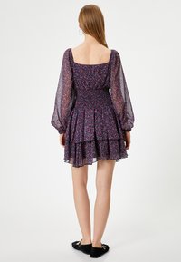 Vestido floral morado con mangas largas transparentes, cuerpo fruncido y falda con volantes en capas. Llevado con zapatos planos negros.