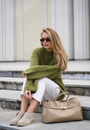 Woman with long blonde hair wearing sunglasses, an oversized green sweater, white pants, and beige shoes sitting on steps next to a beige handbag.