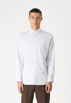 Camicia bianca a maniche lunghe in cotone tessuto con colletto a punta, pattina frontale con bottoni e polsini standard, abbinata a pantaloni marroni.