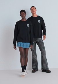 Sorte hettegensere har en sirkulær logo på brystet. Modellen til venstre har på seg et denimskjørt, mens modellen til høyre har på seg bukser med sleng.