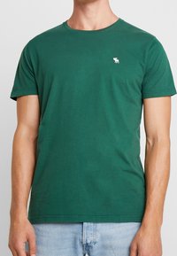 Grünes Baumwoll-T-Shirt mit rundem Halsausschnitt, kurzen Ärmeln und einem kleinen weißen Logo auf der linken Brust. Klassische Passform und glatte Textur.