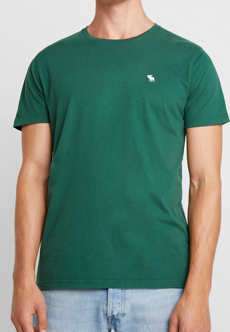 Grünes Baumwoll-T-Shirt mit rundem Halsausschnitt, kurzen Ärmeln und einem kleinen weißen Logo auf der linken Brust. Klassische Passform und glatte Textur.