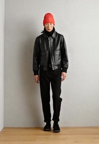 Veste bomber en cuir noir avec deux poches avant, portée sur un col roulé noir et un pantalon noir. Accompagnée d'un bonnet en tricot rouge vif.