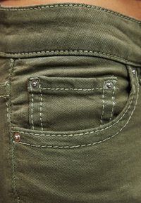 Τζιν παντελόνι σε ελιά πράσινο με πίσω τσέπη με κεντημένες λεπτομέρειες και μεταλλικά κουμπιά. Γενικά ομαλή υφή και εφαρμοστό σχέδιο.