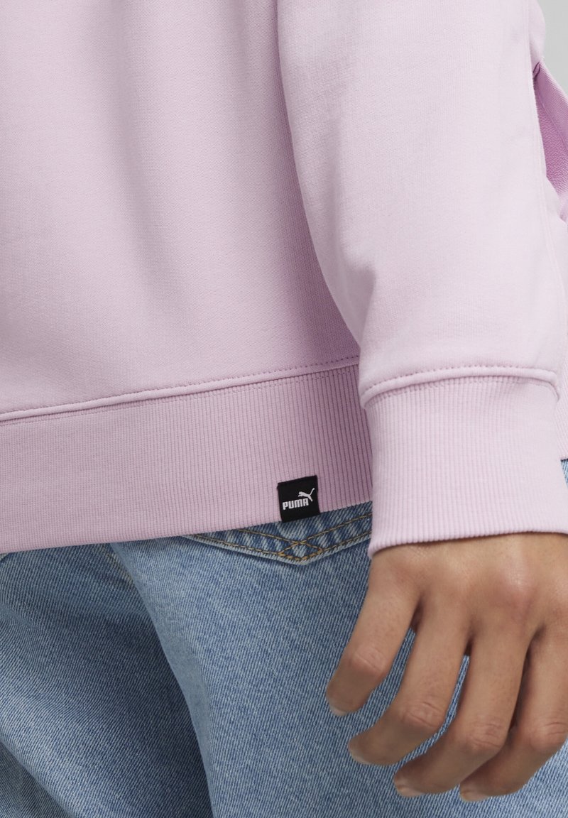 Gros plan d'une personne portant un sweat-shirt rose clair et un jean bleu avec une étiquette visible du logo Puma noir près de la ceinture.