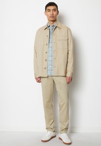 Beige button-up jacket over a light blue plaid shirt, paired with beige trousers and white sneakers. Smooth fabric, casual design.