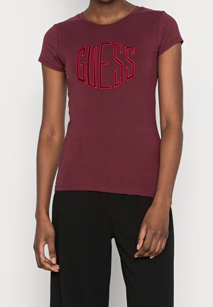 Personne portant une chemise ajustée à manches courtes de couleur bordeaux avec le lettrage "GUESS" en relief et un pantalon noir, se tenant devant un fond uni.