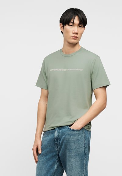 Νεαρός άνδρας με μαύρα μαλλιά, ντυμένος με πράσινο λαχανί T-shirt Emporio Armani και μπλε τζιν, στέκεται με το ένα χέρι στην τσέπη σε απλό φόντο.