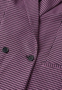 Blazer à motif pied-de-poule violet et noir avec deux boutons noirs et une poche avant visible près du bas à droite.