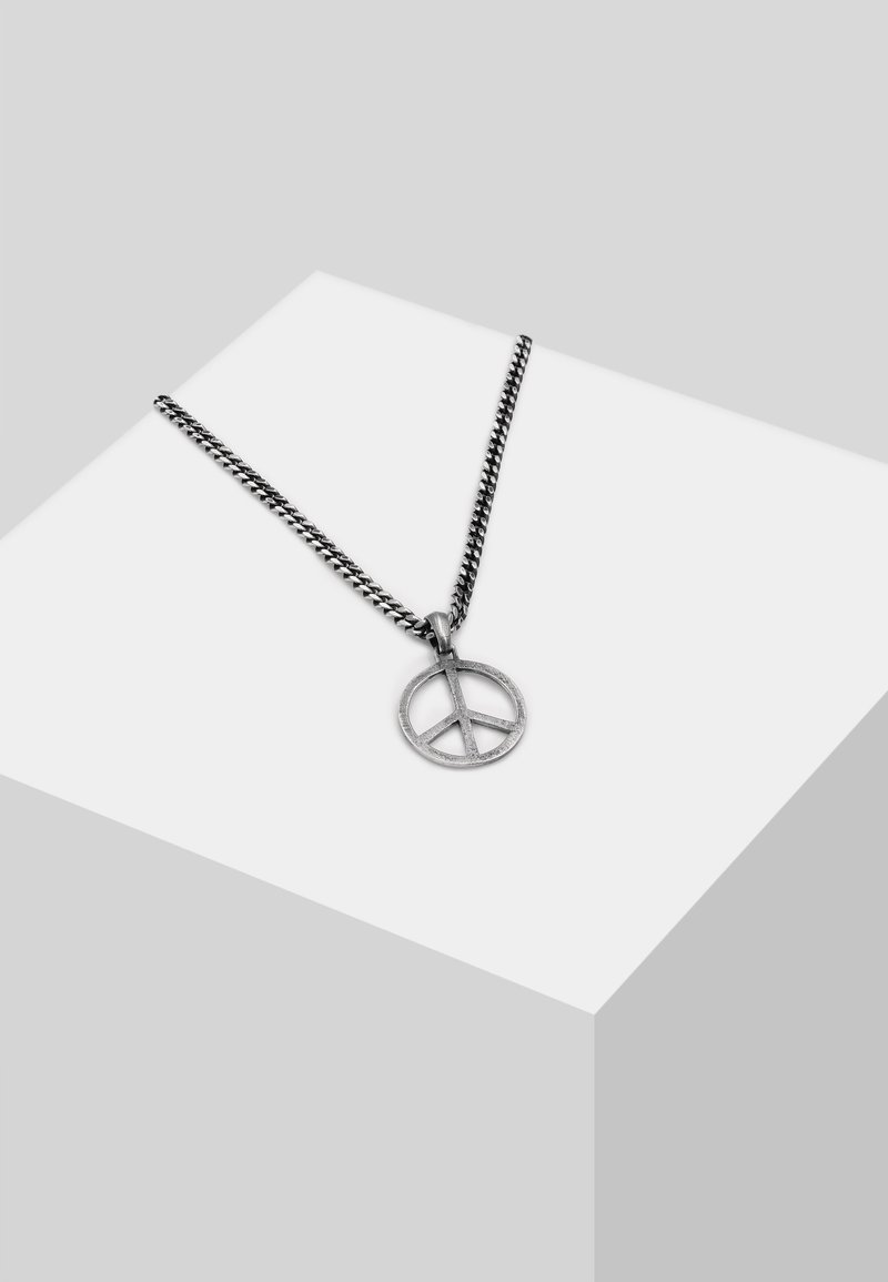 Silverkedja med en cirkulär fredssymbol hängande som har en texturerad yta, placerad mot en minimalistisk bakgrund.