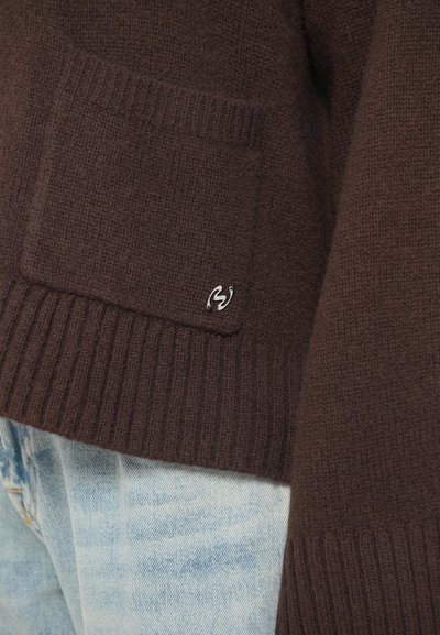 Pulover tricotat de culoare maro, cu o buzunar frontal și un mic insigne argintie, purtat cu blugi din denim de culoare albastru deschis.