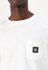 White cotton t-shirt with a round neckline and short sleeves, featuring a patch pocket on the left chest with a black logo label.