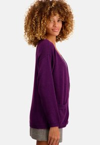 Cardigan en tricot violet avec un col en V, des épaules tombantes, des poches avant et une fermeture à boutons. Matière en mélange de laine texturée.
