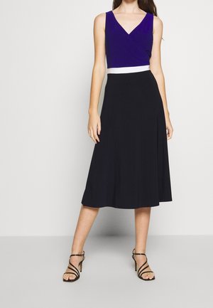 Femme portant un haut violet sans manches rentré dans une jupe midi noire avec une ceinture blanche, associée à des escarpins noirs à brides.
