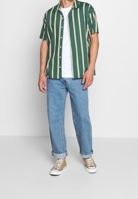 Grön kortärmad skjorta med gula ränder, vit T-shirt under, ljusblå jeans och beige skor med vita sulor.