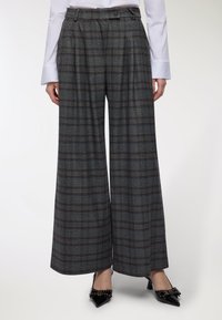 Donna che indossa pantaloni larghi a scacchi grigi con vita alta, abbinati a una camicia bianca a maniche lunghe e tacchi a punta neri con fiocchi.